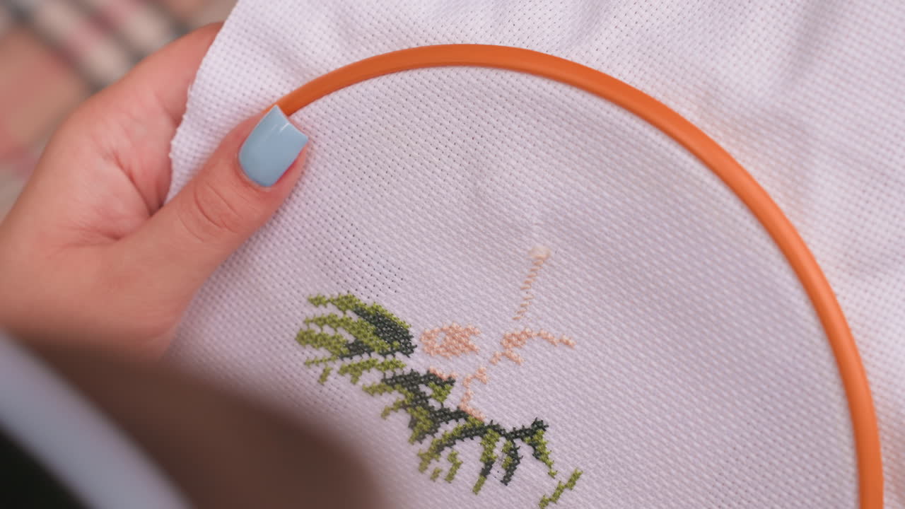 Close up of stylist with painted nails embroidering fabric inside orange hoop, creating detailed tree like cross stitch design with green and beige threads, showing calm focus
