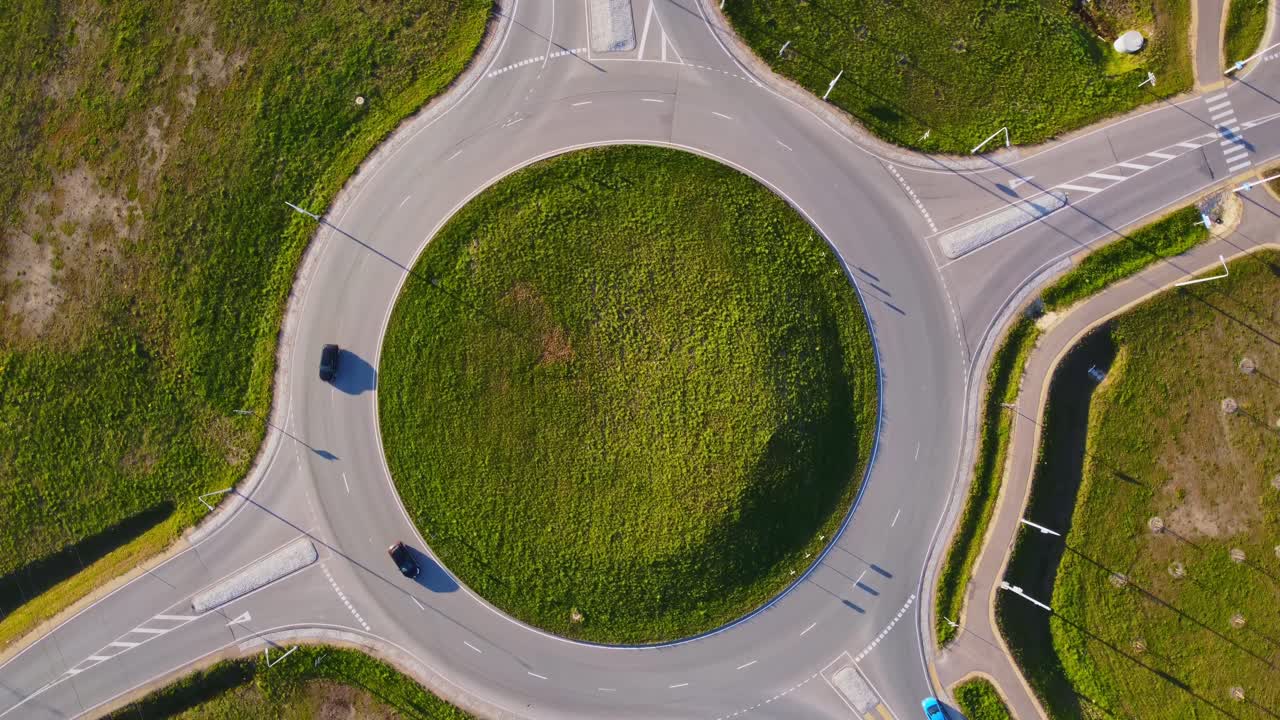 Spinning top down drone shot captures a circular traffic roundabout with grassy central island and two cars navigating the lanes in Katlakalns, Latvia