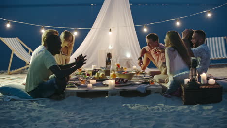 grupo de amigos cenando sentados en la playa, sonriendo y brindando, con una mesa baja y una carpa