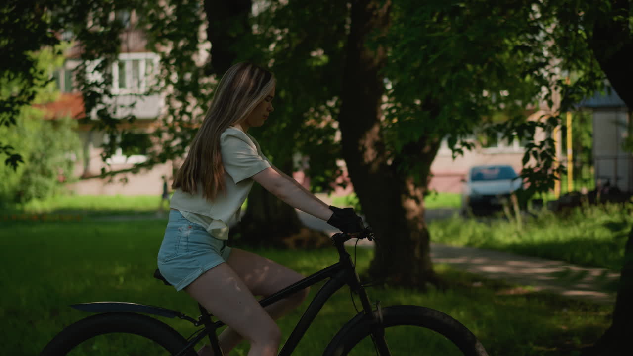 나무 가지로 부분적으로 숨겨진 울창한 초록색 공원을 통해 자전거를 자신감 있게 타고 있는 젊은 여성의 측면 시각, 긴 머리카락이 자유롭게 흐르고, 배경에는 초록색 잎자루와 건물이 있습니다.