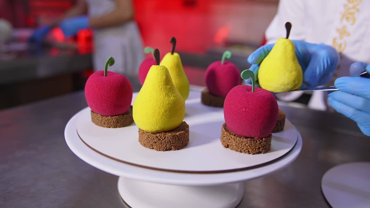 Lovely cakes in the shape of pears and apples on the stand. Confectioner's hands take the pastry and put it on the plate. Close up.