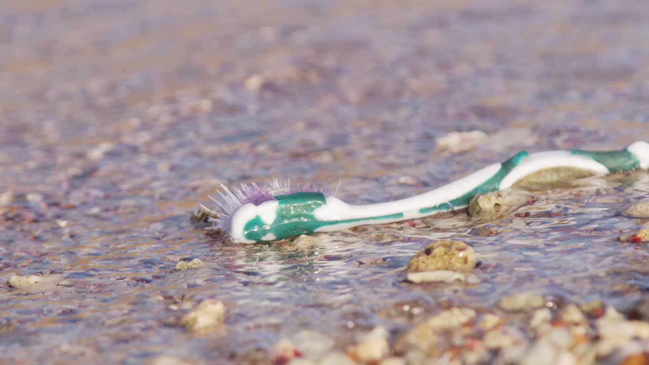 primer plano de un cepillo de dientes viejo de plástico varado en una playa de arena siendo arrastrado por la corriente y las olas