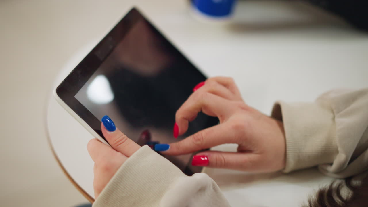 Close up of female hand with brightly painted nails holding tablet and sliding screen while sitting at table indoors, reflecting modern digital lifestyle and casual communication