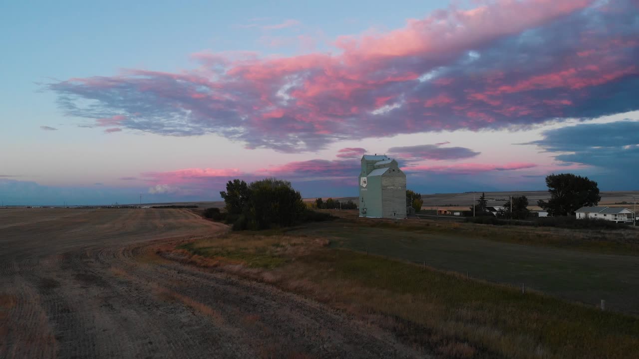 Tomada aérea de drone em movimento frontal de campos agrícolas e um silo de grãos branco em Alberta, Canadá, ao entardecer.