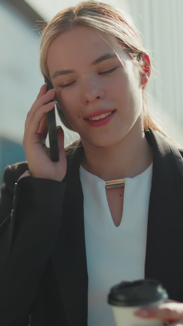 Banker on break taking juice while on phone call, walking past glass facade building under bright daylight, reflecting surrounding cityscape with soft sunlight illuminating her