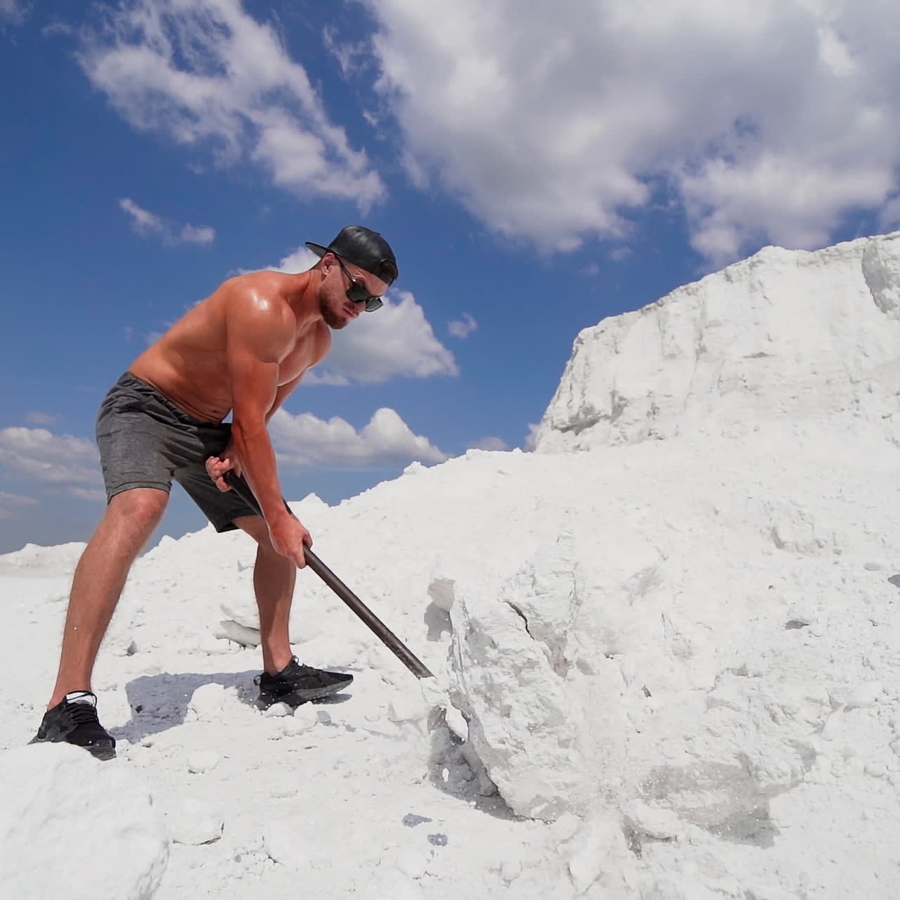 Strong man with steel hammer works on white hill under blue sky. Sporty male in shorts breaks big stone by heavy hammer outdoors.