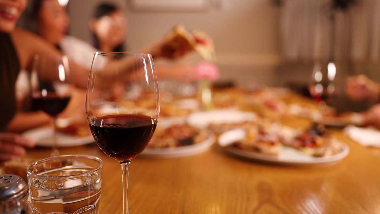 Primo piano di un bicchiere di vino rosso con un gruppo sfocato che condivide una pizza in un'illuminazione calda del ristorante.