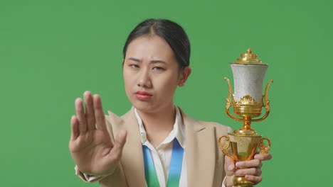 close up of asian business woman in a suit with a gold medal and trophy disapproving with no hand sign and smiling to camera on green screen background in the studio