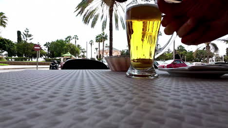 primer plano de una mano recogiendo un vaso de cerveza en una mesa con una calle alineada con palmeras en el fondo