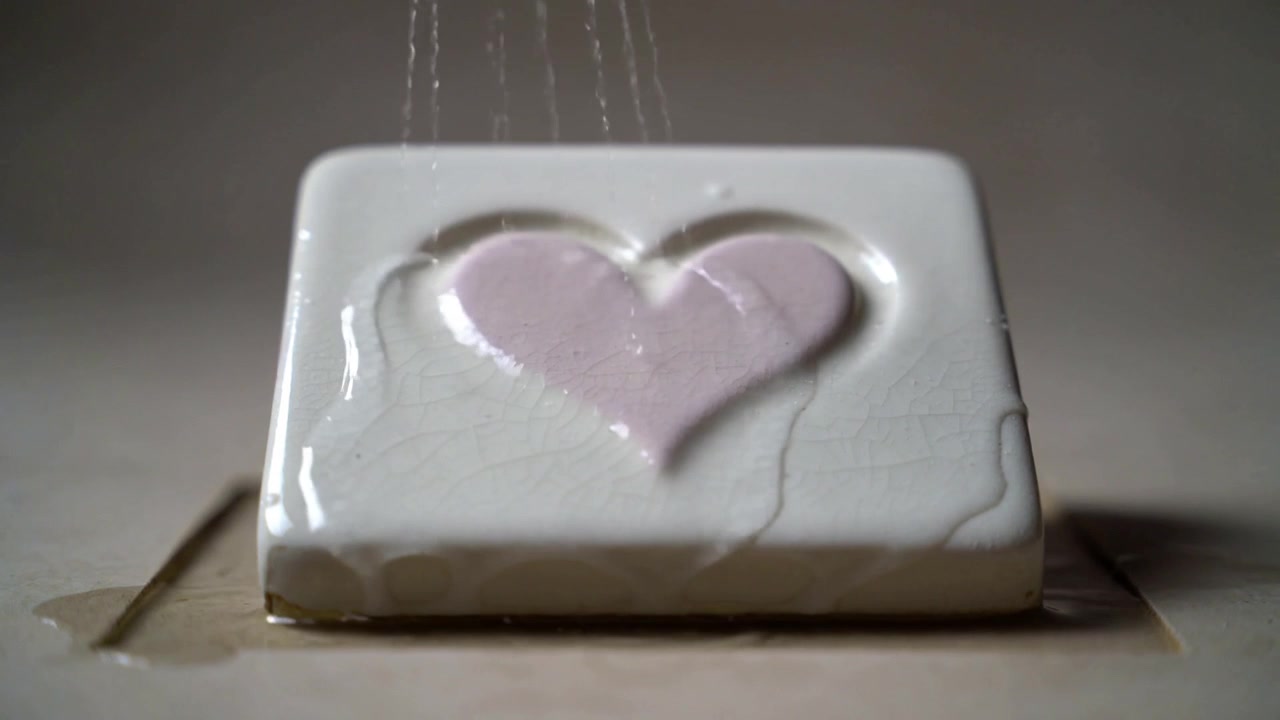 Getti d'acqua che cadono colpiscono una piastrella smaltata con un cuore rosa pallido in rilievo su un vassoio, mostrando delle screpolature.