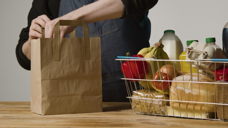 Studio Shot Of Shop Worker Packing Basic Food Items In Supermarket Wire Shopping Basket Into Paper Bag 3
