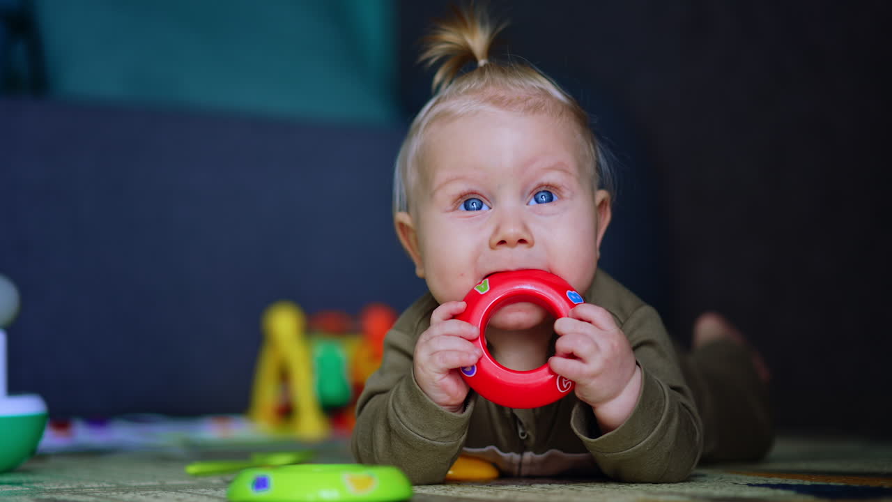 Adorable chubby infant lies on the floor on belly. Lovely baby chews the toy, looks up and smiles. Close up.