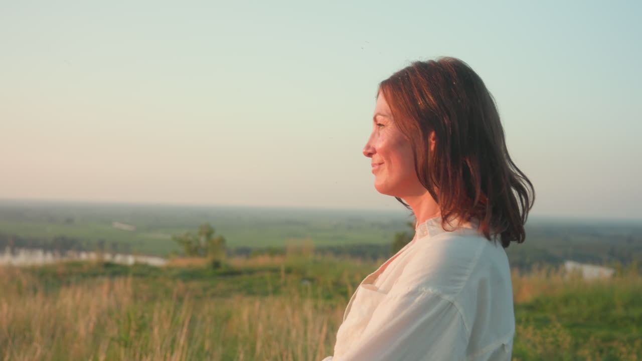 Side view of woman smiling as she shapes rectangle with hands against rural backdrop, sunlight illuminating hair and blouse sleeve, evening light casting soft glow on joyful face and gesture