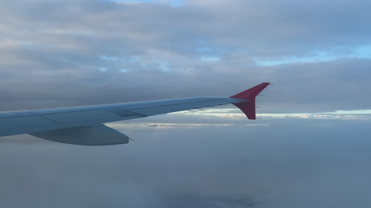 Kommersiell flyvinge sett fra et passasjervindu som flyr over et tett skylag under en stemningsfull overskyet himmel. Subtilt lys bryter gjennom horisonten, og skaper en rolig og atmosfærisk reisesscene.