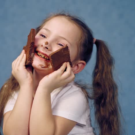 little girl with dirty face eats bar of chocolate. funny child with two ponytails is biting tasty black chocolate on blue studio background.