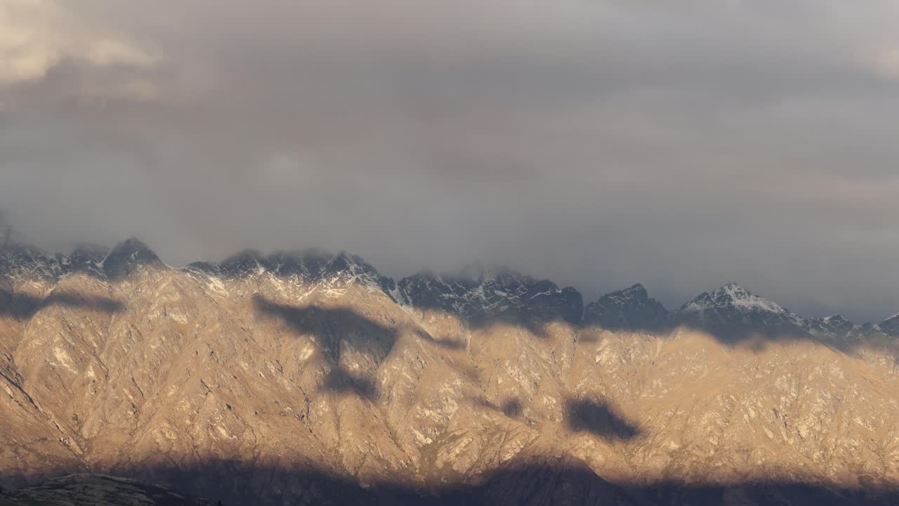 Clouds move over a rugged mountain range, casting dynamic shadows. The scene captures changing light and atmospheric conditions