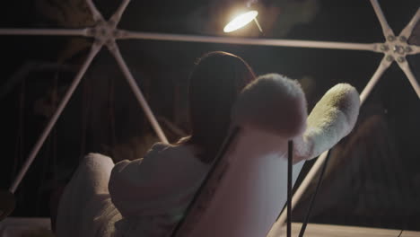 woman with dark hair in soft bathrobe looks out huge window. lady relaxes in comfortable armchair after arduous workday on balcony lit by light of lantern