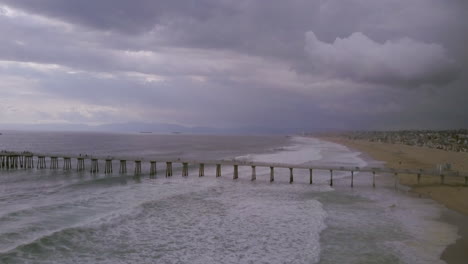 Heavy-summer-storm-waves-rolling-past-the-Manhattan-Beach-Pier-in-California,-AERIAL