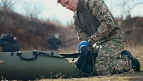 View-from-the-side-of-a-confident-young-male-army-medic-in-a-camouflage-green-uniform-strapping-and-stabilizing-an-unconscious-man-soldier-on-a-medical-army-stretcher-at-an-army-training-ground