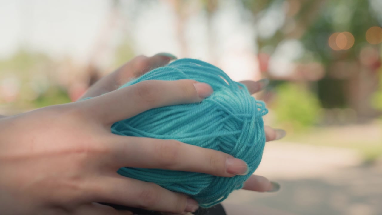 Close up of hands holding and manipulating ball of turquoise yarn in outdoor setting with soft focus background. person fingers gently grasp textured thread showcasing its color and material