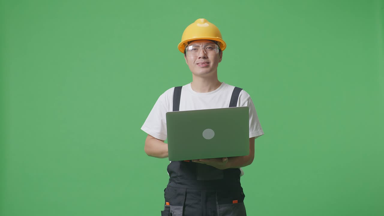 Asian Man Worker Wearing Goggles And Safety Helmet Using A Laptop And Smiling While Standing In The Green Screen Background Studio