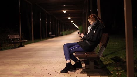 Mujer-Joven-Sola-Mirando-Su-Teléfono-Inteligente-Por-La-Noche-En-Un-Banco-Del-Parque