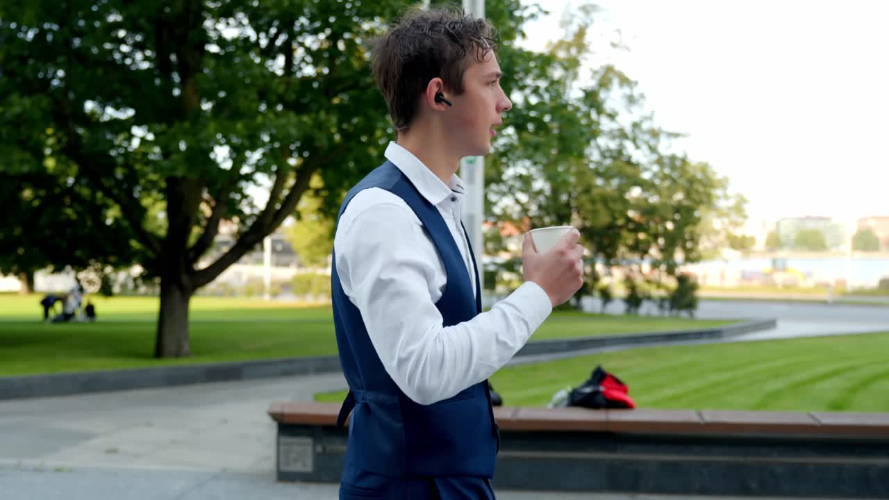 Young businessman walking, wearing a blue vest and white shirt, holding coffee, using a wireless earbud, enjoying a break outdoors in a green city park, slow motion shot