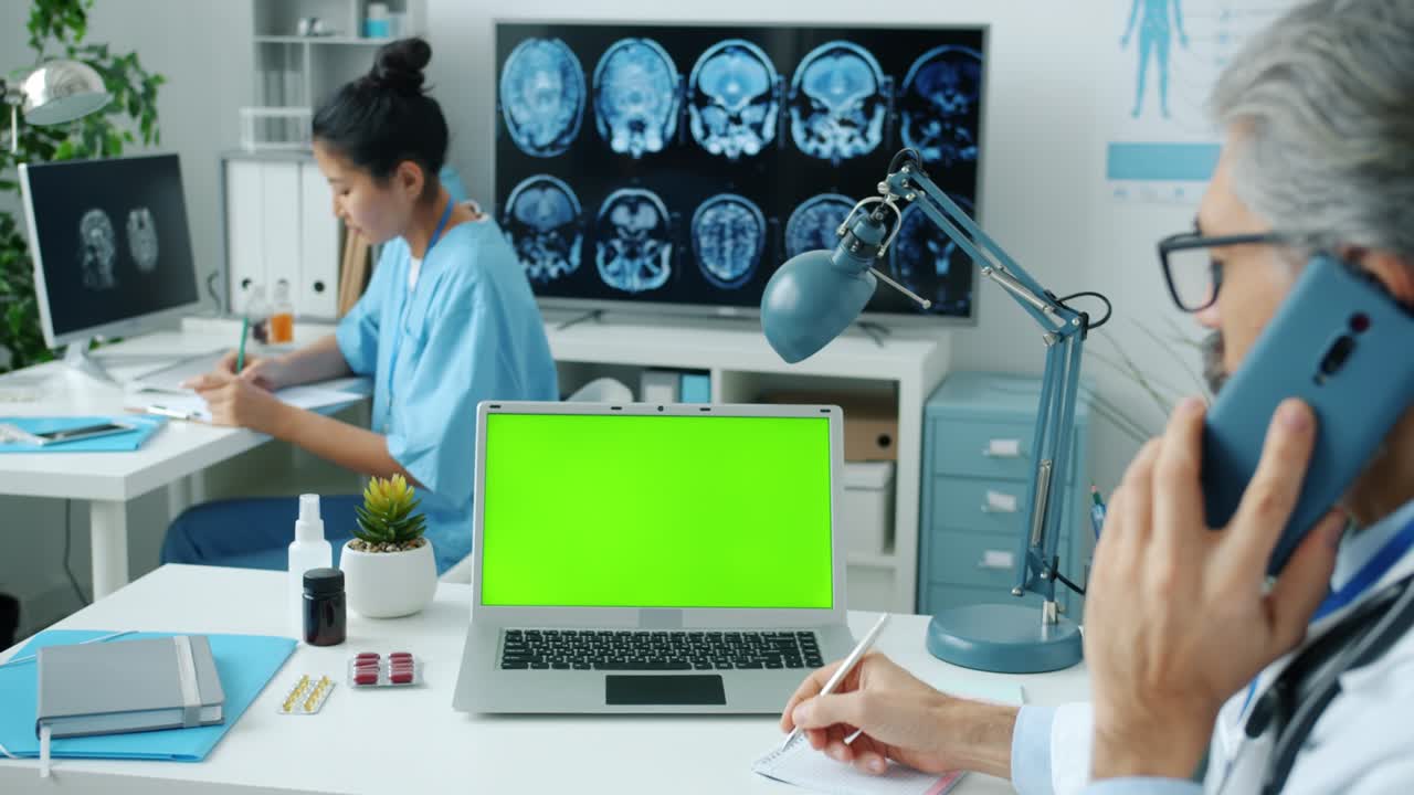 el médico hablando en el teléfono móvil y mirando la pantalla verde de la computadora portátil de clave de croma mientras la enfermera escribe en segundo plano.