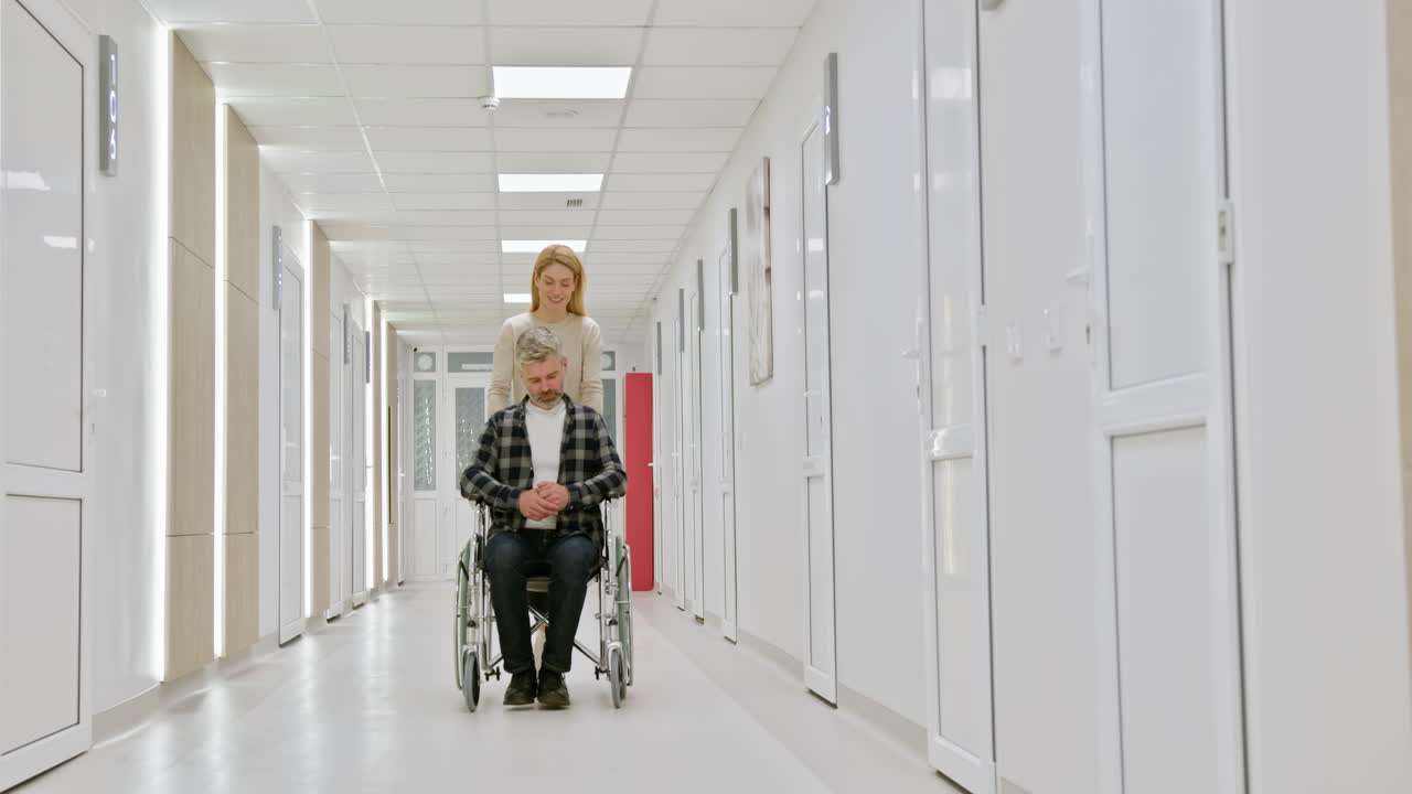 hombre en silla de ruedas siendo empujado por una mujer en el pasillo del hospital