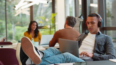 young businessman with feet on desk in office wearing wireless headphones  greeting colleague