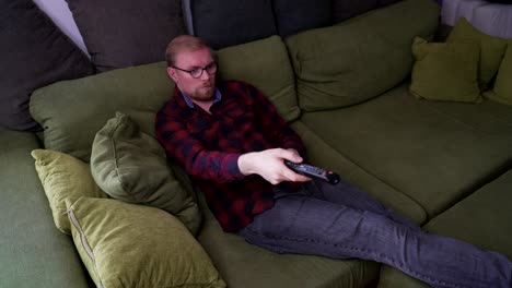 young man lays on sofa, uses tv remote to switch channels, medium shot