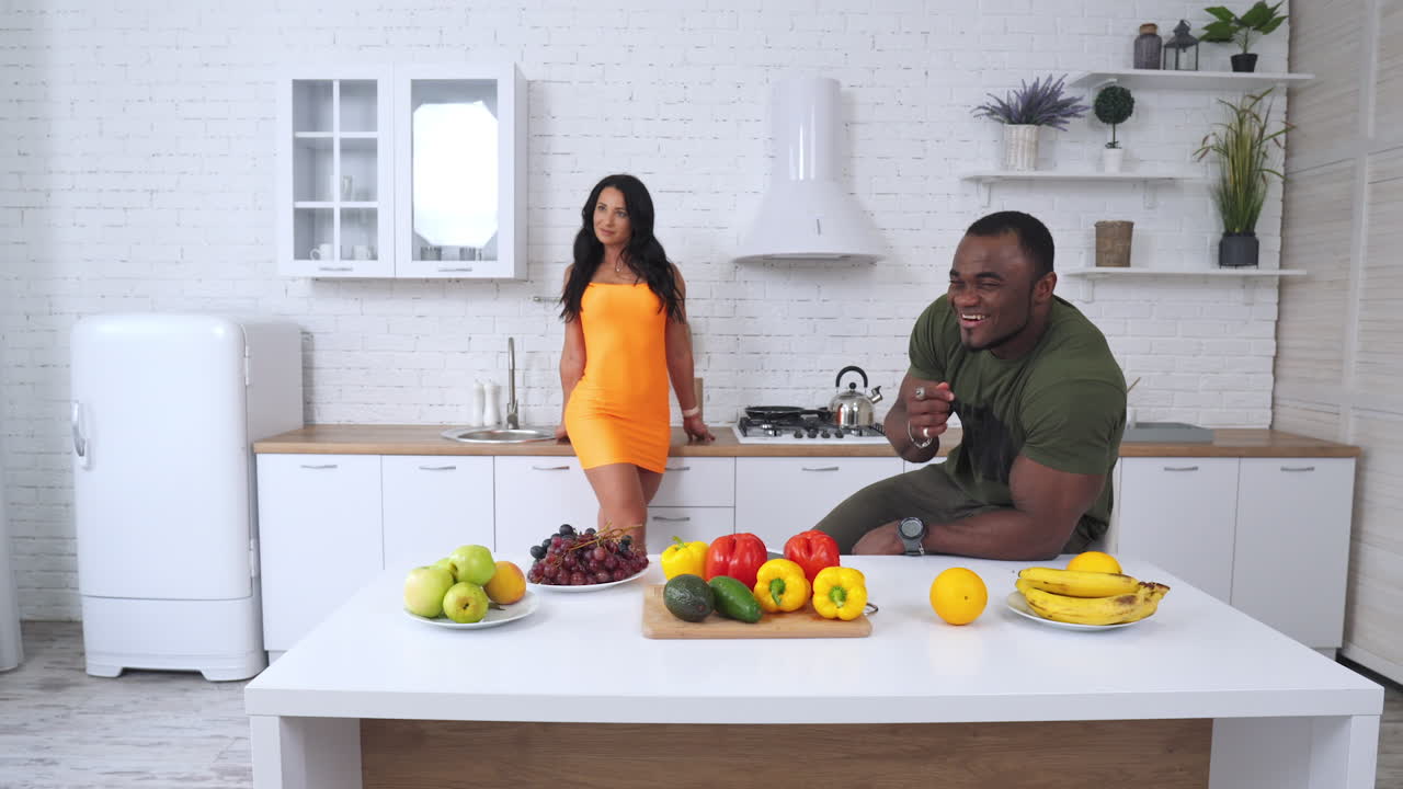 Adult man with big muscles sitting at the table and eating grapes. Lady in orange mini dress standing behind. Male beckoning to female and she comes up to a table with fruit and vegetables.