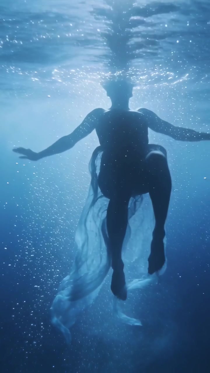 Video verticale: Una donna nuota verso l'alto in acqua blu con un abito fluttuante, reagendo alla luce superficiale.