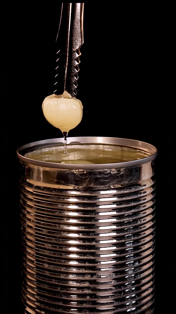 Pinze metalliche afferrano e sollevano un frutto di longan sbucciato da una lattina aperta piena di sciroppo, sotto un'illuminazione drammatica da studio su sfondo nero