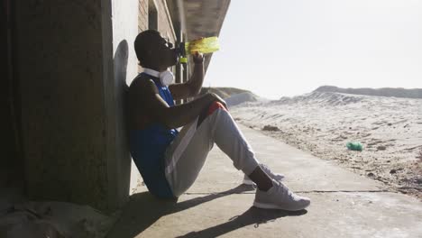 hombre afroamericano sentado y bebiendo de una botella de agua tomando un descanso en el ejercicio al aire libre