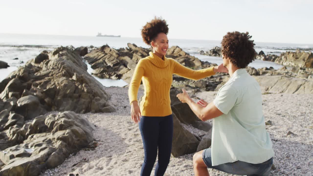 African american man putting a ring on his girlfriend's finger on the rocks near the sea