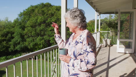 caucasian senior woman holding coffee cup waving while standing in the balcony at home