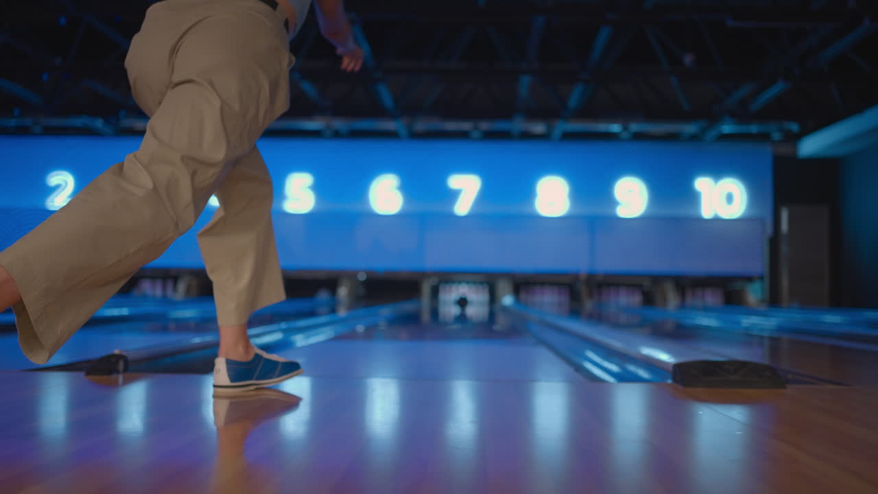 Kamera sleduje ženu, která hází bowlingovou kouli na hrací dráhu a radostně skáče z poražených kuželek. Muž hraje bowling ve zpomaleném záběru.