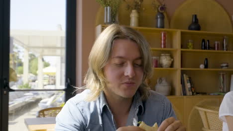 Front-View-Of-Blond-Man-Eating-Pizza-Sitting-At-A-Restaurant-Table-With-His-Friends