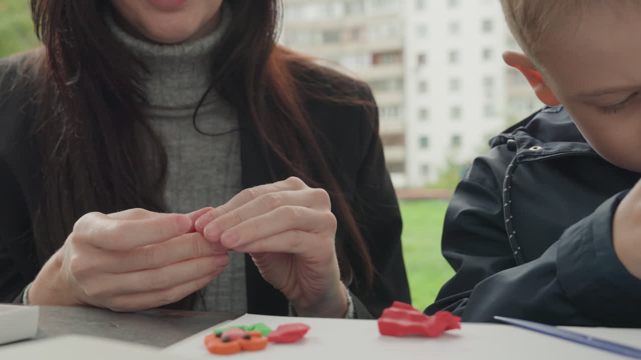 Madre e figlio caucasici all'aperto che creano con l'argilla su un tavolo, sullo sfondo un condominio, la madre che guida le mani del bambino, forme modellate rosse, istruzione e insegnamento pazienti, un momento di apprendimento giocoso.