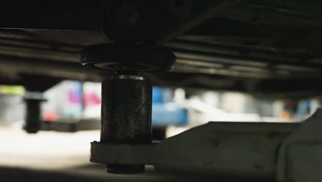 close-up of car lift positioned under vehicle in auto repair shop, focusing on mechanical details with subtle movement in blurred background