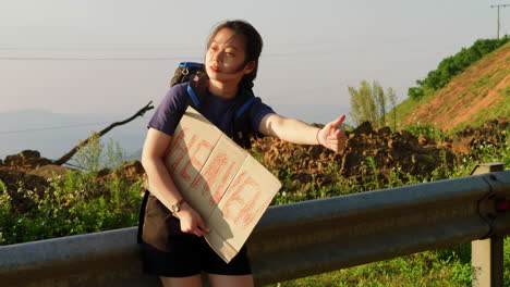 slow-motion-of-young-female-asiatic-traveller-hitchhiking-with-thumbs-up-and-a-sign-to-heaven-on-cardboard