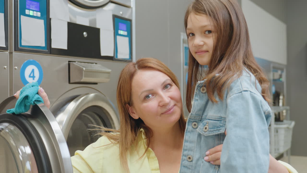 Laundry worker crouches with towel, hugs little girl beside stainless washer in laundromat, both smiling during cleaning break, warm family moment, teamwork, care, hygiene focus indoors