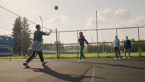 lady practicing volleyball with coach, hitting ball over net while two other teammates watch, demonstrating team training session in outdoor court, with clear sky and surrounding greenery