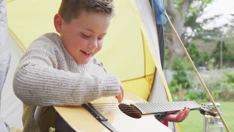 Feliz-Niño-Caucásico-Sentado-En-Una-Tienda-De-Campaña-En-El-Jardín-Y-Tocando-La-Guitarra