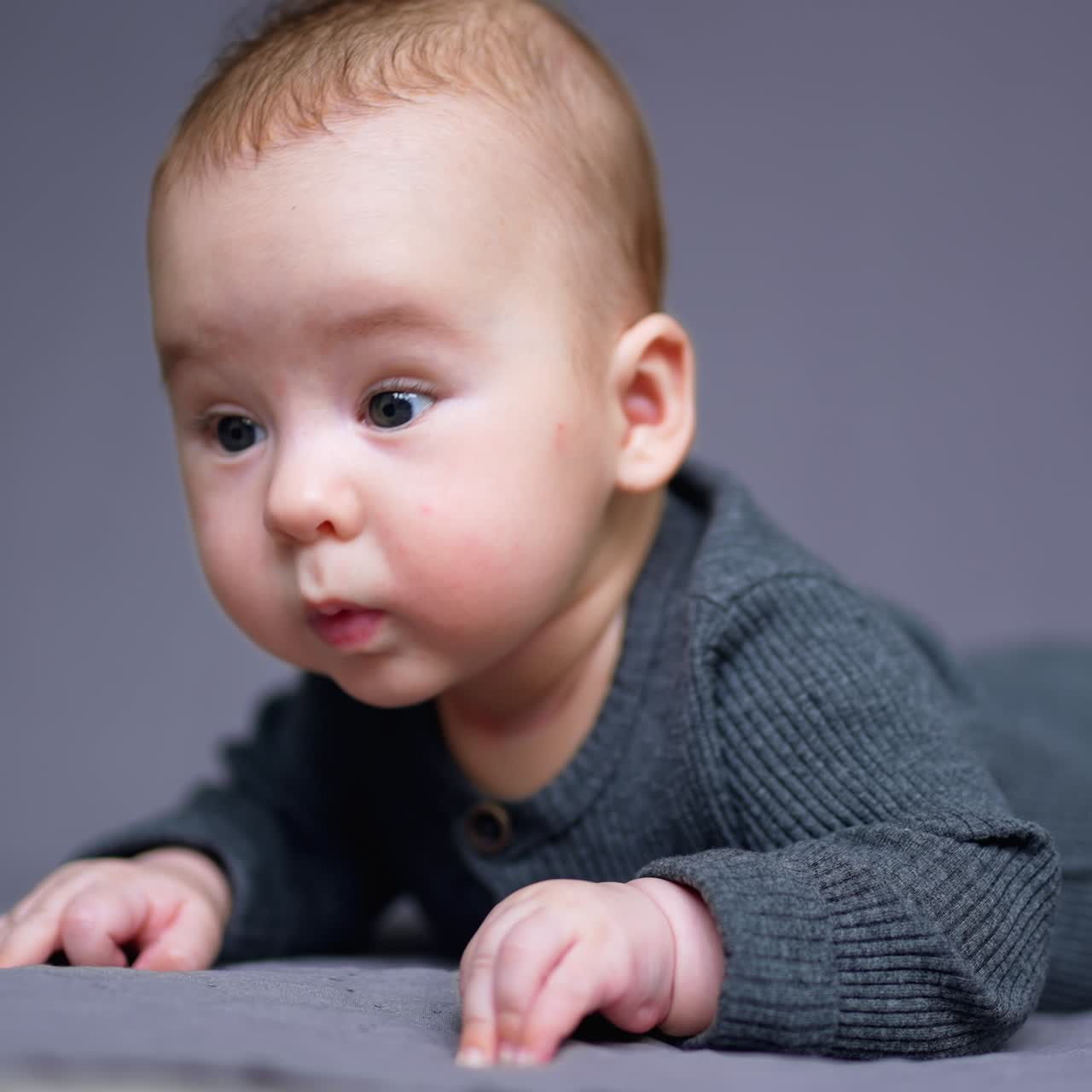 Little cute baby boy in grey outfit rests on his belly. Lovely child looks sideways with interest. Grey blurred backdrop