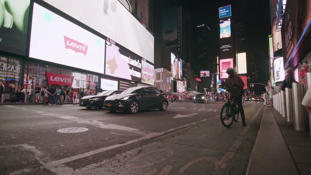A busy night in Times Square with neon billboards, street vendors, cars, and a cyclist