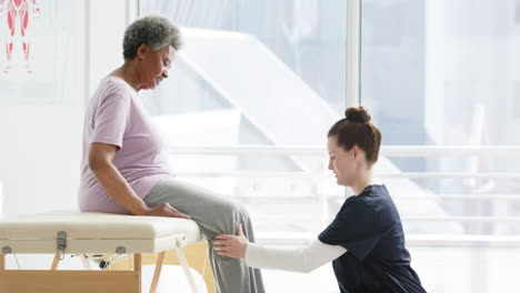 diverse senior woman exercising leg and female doctor advising in hospital, slow motion