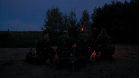 a team of men in camouflage clothing is sitting on a hill in the field at night, warming themselves by a lit campfire, frying sausages for dinner, a manly outing, relaxing after work, service, hunting