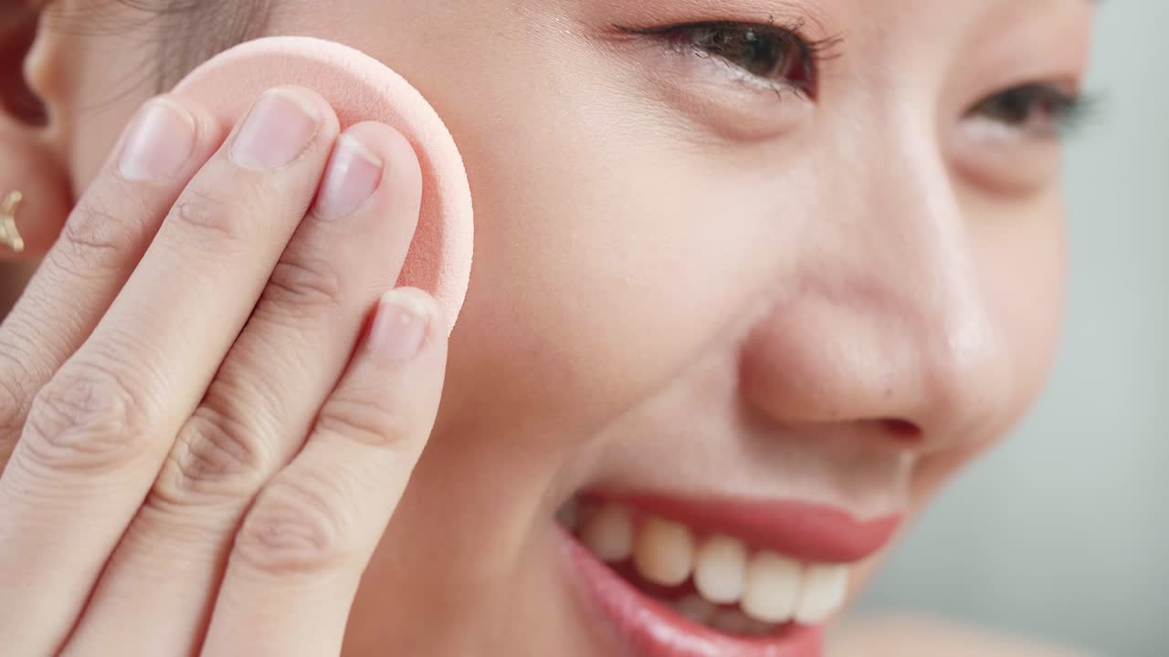 아름다운 젊은 아시아 여성이 웰니스 피부 관리를 위해 파우더 얼굴을 적용하고 집에서 메이크업하는 동안 미소 짓고 있습니다.